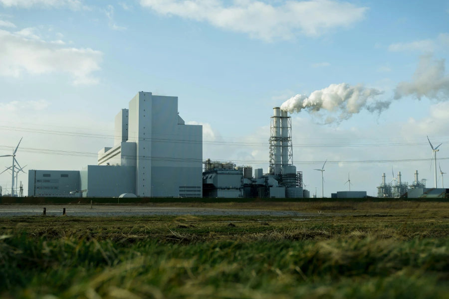 Ett stort, modernt industriverk eller kraftverk med en rykande skorsten, omgivet av vindkraftverk under en delvis molnig himmel. I förgrunden syns ett grönt gräsfält.